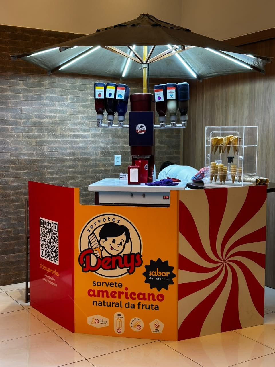 Quiosque Denys Sorvetes em shopping, com braço de sabores e guarda-sol iluminado